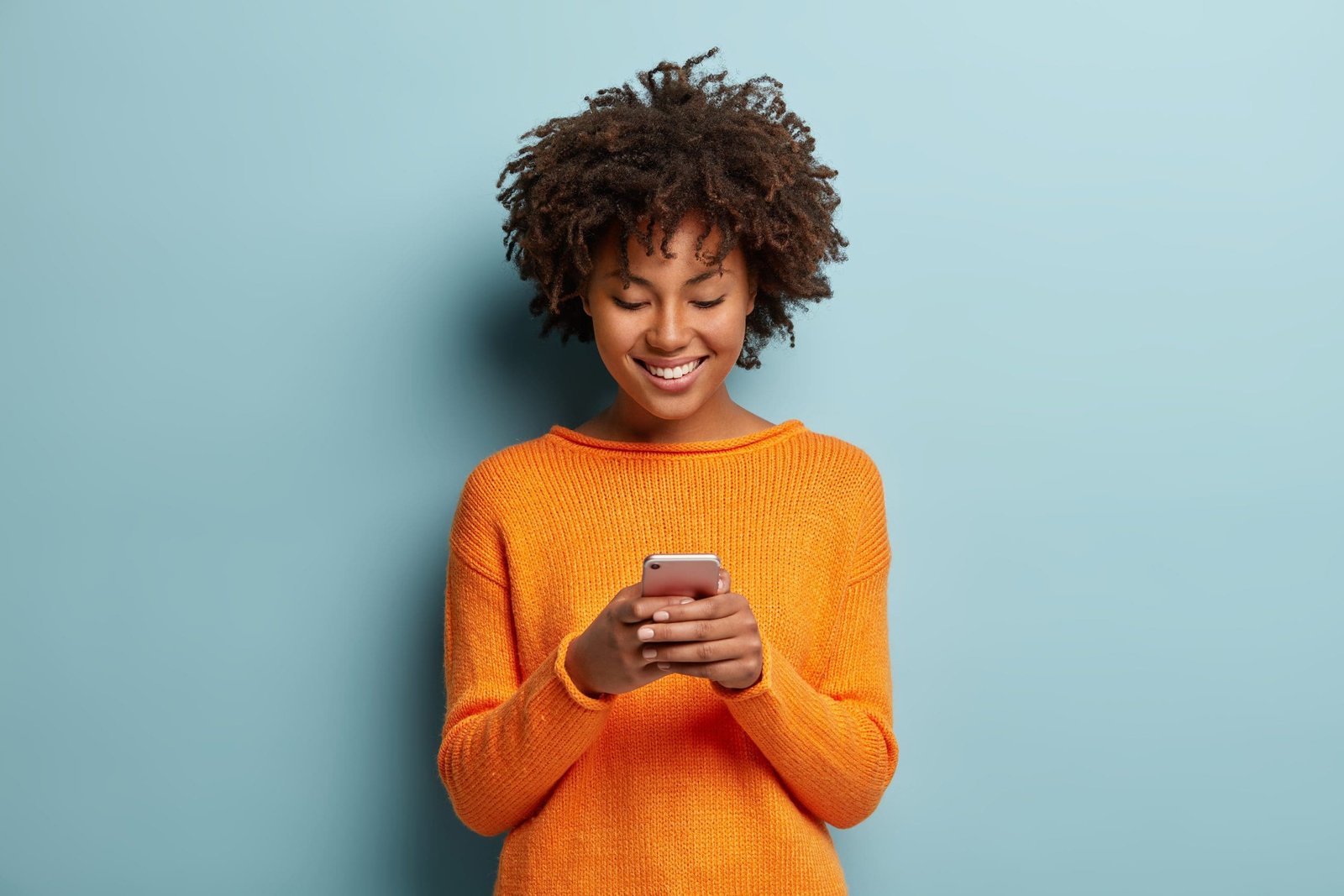 Satisfied hipster girl with Afro haircut, types text message on cell phone, enjoys online communication, types feedback, wears orange jumper, isolated on blue studio wall. Technology concept